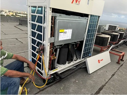 Two people beside an open outdoor unit and connections on a rooftop