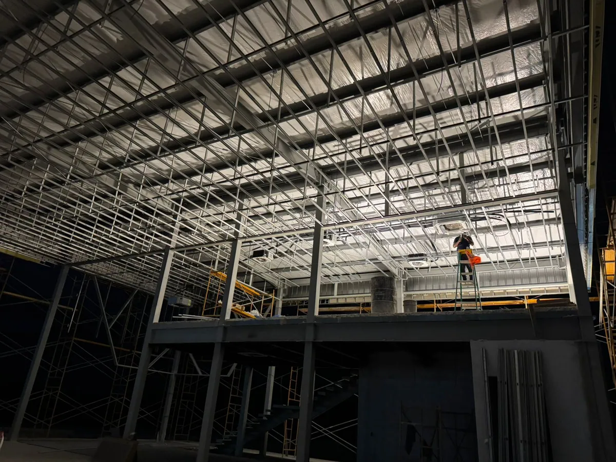 Wide interior with exposed roof framing, suspended equipment, and a person in the distance